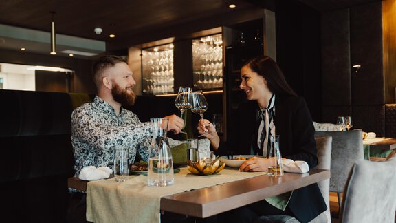 Ein Paar sitzt in einem eleganten Restaurant und stößt mit Weingläsern an. Beide lächeln und genießen ihr gemeinsames Essen. - VIER - Studios & Restaurant - Längenfeld