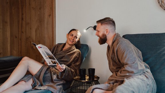 Ein Paar entspannt in Bademänteln in einem gemütlichen Raum. Sie sitzen auf Liegen und unterhalten sich, während sie eine Zeitschrift lesen. - VIER - Studios & Restaurant - Längenfeld