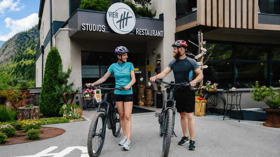 Ein Paar steht mit Fahrrädern vor einem modernen Gebäude, das ein Restaurant und Studios beherbergt. Die Umgebung ist grün und die Atmosphäre wirkt einladend. - VIER - Studios & Restaurant - Längenfeld