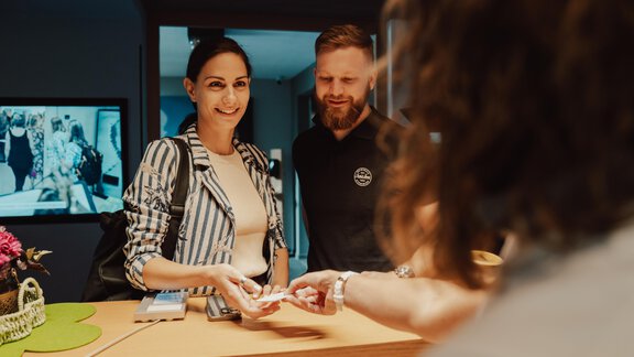 Ein Empfangstresen mit einer lächelnden Frau und einem Mann, die miteinander sprechen. Im Hintergrund ist ein Bildschirm mit Bildern zu sehen. - VIER - Studios & Restaurant - Längenfeld
