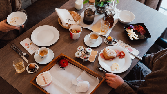 Ein gedeckter Tisch mit verschiedenen Frühstücksgerichten, darunter Gebäck, Obst und Kaffee. Blumen und Milchkrüge ergänzen das gemütliche Ambiente. - VIER - Studios & Restaurant - Längenfeld
