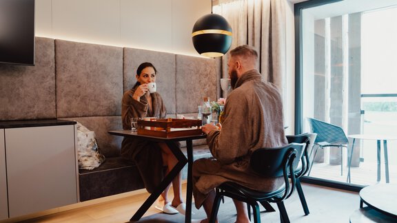 Ein Paar in Bademänteln sitzt an einem Tisch und genießt eine Tasse Kaffee. Der Raum ist modern eingerichtet mit einem großen Fenster und gemütlicher Atmosphäre. - VIER - Studios & Restaurant - Längenfeld