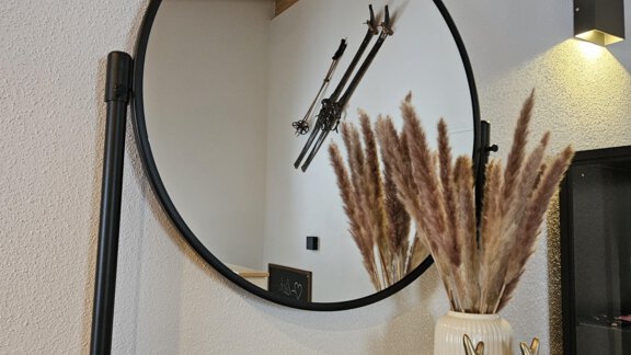 Ein runder Spiegel an einer Wand mit Holzdecke. Darunter steht eine dekorative Vase mit Trockenblumen und ein goldenes Rehfigur auf einem runden Tablett. - Apart Alpenflair - Niederthai