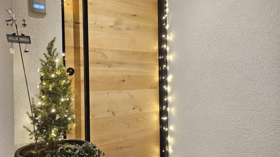 An inviting front door made of warm wood with festive lighting. Next to the door stands a small Christmas tree in a pot. - Apart Alpenflair - Niederthai