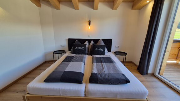 Ein modernes Schlafzimmer mit einem Doppelbett und angenehmer Beleuchtung. Große Fenster bieten Blick nach draußen und sorgen für Helligkeit im Raum. Für Unterhaltung sorgt ein FlatTV. - Apart Alpenflair - Niederthai