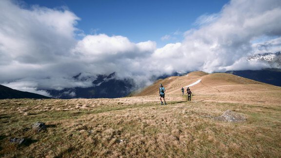 Zwei Wanderer gehen über eine grüne Berglandschaft unter einem bewölkten Himmel. Im Hintergrund sind majestätische Berge und Wolken zu sehen. - Apart Alpenflair - Niederthai