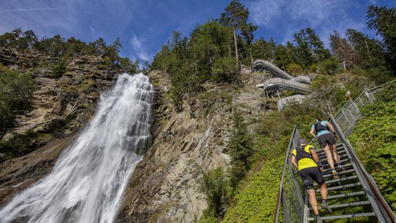 Eine Gruppe von Menschen steigt eine Treppe hinauf, die zu einem Wasserfall führt. Im Hintergrund sieht man die hübsche Natur mit Bäumen und einem blauen Himmel. - Apart Alpenflair - Niederthai