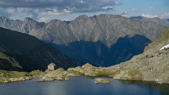 Ein idyllischer Bergsee umgeben von beeindruckenden Berggipfeln. Eine alte Hütte liegt malerisch am Ufer. - Apart Alpenflair - Niederthai