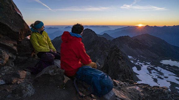 Zwei Wanderer sitzen auf einem Berg und genießen den Sonnenuntergang. Die Landschaft zeigt majestätische Berge und schneebedeckte Flächen im Hintergrund. - Apart Alpenflair - Niederthai