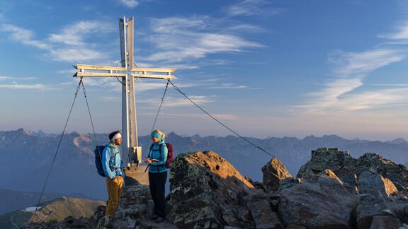 Zwei Wanderer stehen am Gipfel eines Berges vor einem großen Kreuz. Im Hintergrund sind beeindruckende Berggipfel und ein schöner Himmel zu sehen. - Apart Alpenflair - Niederthai