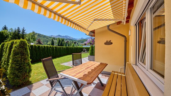 A cozy terrace with a wooden table and chairs. In the background, green plants and the mountains are visible. - Appartement Lea - Niederthai