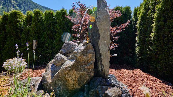 A beautiful garden with a waterfall made of stones.  
Surrounded by green hedges and colorful flowers under a blue sky. - Appartement Lea - Niederthai