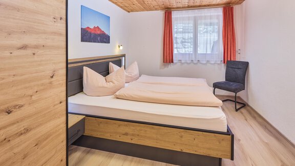 A cozy bedroom with a double bed and two pillows. There is an armchair and a window with curtains that provides natural light. - Appartement Lea - Niederthai