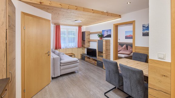 A cozy living kitchen with a modern sofa, a dining table, and wooden shelves. The walls are light, and there are large windows with red curtains. - Appartement Lea - Niederthai