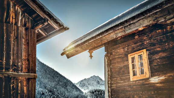 Two old wooden buildings come together, surrounded by snow-covered mountains. The sun is shining and gently illuminating the scene. - Appartement Lea - Niederthai