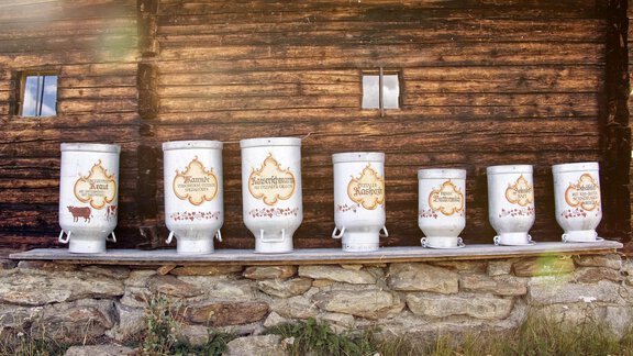 A row of decorative milk cans stands against a wooden wall. The cans are in various sizes and adorned with colorful labels. - Appartement Lea - Niederthai