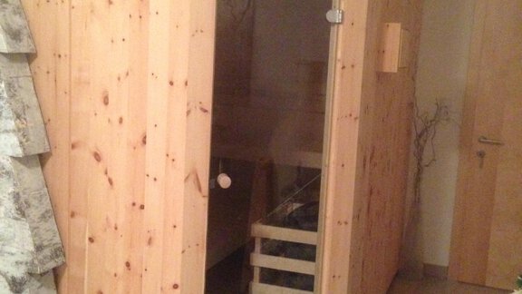 A wooden sauna cabin in a modern room. The walls are made of light wood, and the door is made of glass. - Delano Appartements - Niederthai