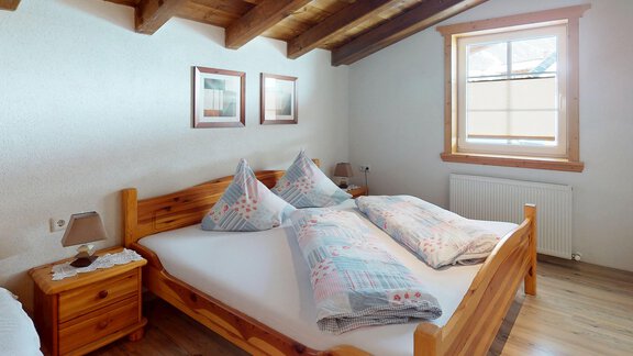 Ein gemütliches Schlafzimmer mit einem Holzbett und zwei großen Kissen. Die Wände sind hell gestrichen, und es gibt ein Fenster mit viel Tageslicht. - Gipfelblick Appartements - Niederthai