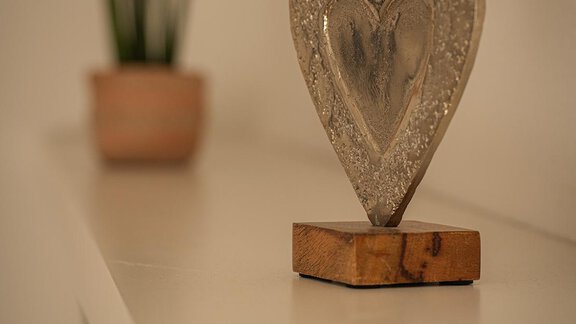 Ein dekoratives Herz aus Stein steht auf einem weißen Regal. Im Hintergrund sind zwei kleine Pflanzen in Töpfen zu sehen. - Sonnenhof - Niederthai