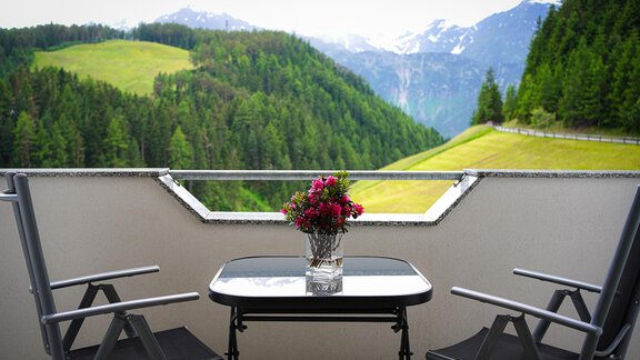 Ein Balkon mit Blick auf grüne Hügel und Berge im Hintergrund. Ein Tisch mit einer Vase voller Blumen und zwei Stühlen lädt zum Entspannen ein. - Sonnenhof - Niederthai