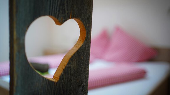 Ein Holzschrank mit einem herzförmigen Ausschnitt. Im Hintergrund sind Kissen mit kariertem Muster zu sehen. - Sonnenhof - Niederthai