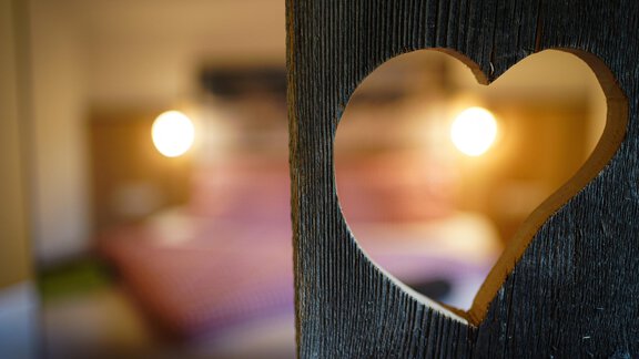 Ein herzförmiger Ausschnitt in einem Holztür zeigt einen gemütlichen Schlafraum. Weiche Beleuchtung und ein einladendes Bett schaffen eine romantische Atmosphäre. - Sonnenhof - Niederthai