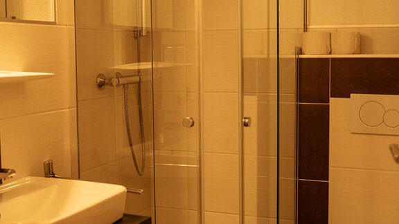 Ein modernes Badezimmer mit einer gläsernen Dusche und einem Waschbecken. Die Wände sind hell gefliest und es gibt eine Toilette im Hintergrund. - Sonnenhof - Niederthai