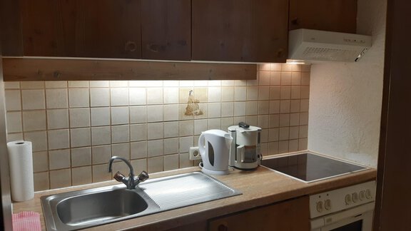 Eine moderne Küche mit Holzschränken und heller Beleuchtung. Zu sehen sind eine Spüle, ein Wasserkocher und ein Kochfeld. - Tirolerhof - Niederthai