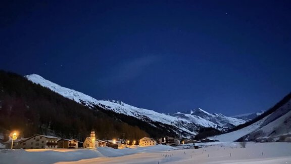 A snow-covered landscape at night with snowy mountains. In the foreground, some buildings and a church are illuminated. - Appartementhaus Schalfblick - Obergurgl 