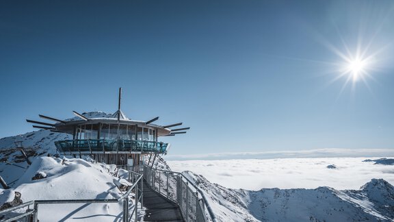 An impressive mountain landscape with snow-capped peaks and a clear blue sky. In the foreground, there is an observation platform that offers a wide view of the clouds below. - Appartementhaus Schalfblick - Obergurgl 