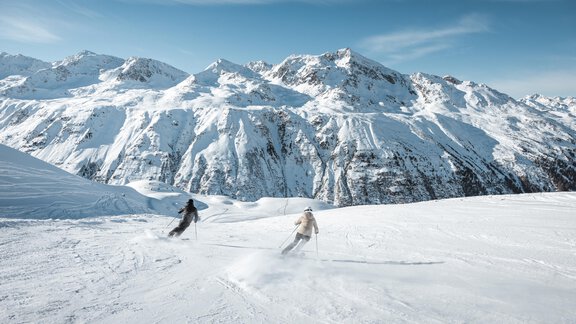 Two people are skiing on a snow-covered slope. In the background, there are impressive, snow-covered mountains and a clear blue sky. - Appartementhaus Schalfblick - Obergurgl 