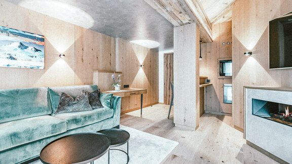 A modern living room with a cozy sofa and a table. The walls are made of wood and there is a small kitchen in the background. - Austria Gourmet & Wine Hotel - Obergurgl 