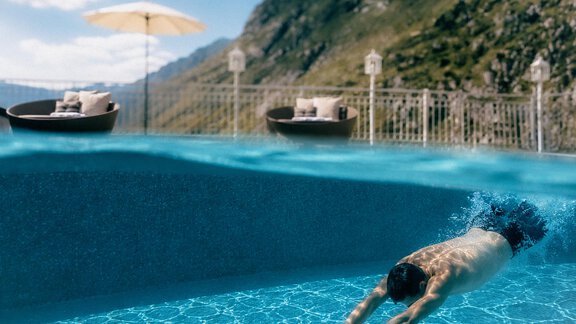 A man dives into a clear, blue swimming pool with a view of mountains. In the background, elegant lounge chairs and a sun umbrella are visible. - Austria Gourmet & Wine Hotel - Obergurgl 