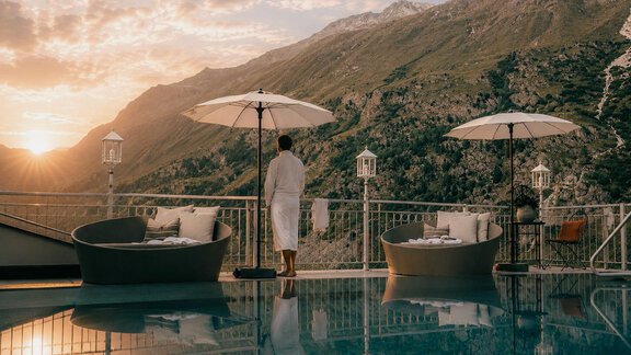 A beautiful sunset over the mountains with a pool in the foreground. A person in a bathrobe stands at the edge of the pool and enjoys the view. - Austria Gourmet & Wine Hotel - Obergurgl 
