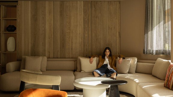 A modern living room with a large, comfortable sofa and a round table. A person is sitting relaxed in the space, surrounded by warm colors and natural light. - Austria Gourmet & Wine Hotel - Obergurgl 