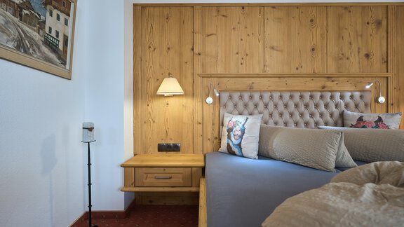 A cozy bedroom with wooden walls and a padded headboard. Next to the bed is a nightstand with a lamp. - Forest Chalet - Obergurgl 