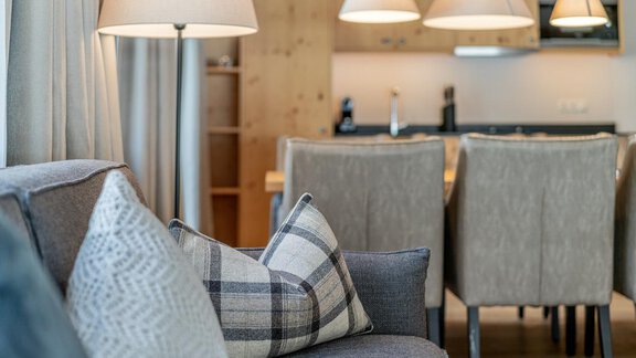 A cozy living room with a gray sofa and decorative cushions. In the background, a dining table and modern lamps are visible. - Gletscherblick - Serviced Apartments - Obergurgl 