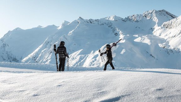 Two skiers are standing on a snow-covered mountain hut. In the background, majestic mountains are visible under a clear blue sky. - Gletscherblick - Serviced Apartments - Obergurgl 