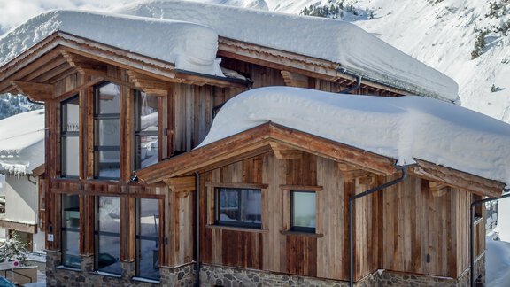 A modern wooden house in the mountains, surrounded by snow. The large window front offers a view of the alpine landscape. - Gletscherblick - Serviced Apartments - Obergurgl 
