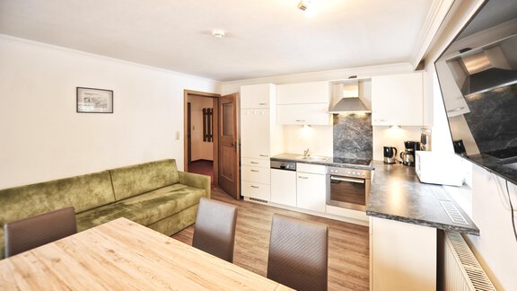 A modern kitchen with white furniture and an open living area. A dining area with a wooden table and chairs is also visible. - Haus Timmelseck - Obergurgl 