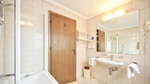 A bright bathroom with a bathtub, a sink, and a large mirror. The walls are tiled and there is a wooden chair as well as towels. - Haus Timmelseck - Obergurgl 