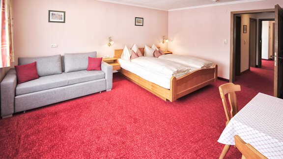 A cozy hotel room with a double bed and a gray couch. The floor is covered with a red carpet and there is a table with chairs. - Haus Timmelseck - Obergurgl 