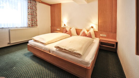 A bright bedroom with a large double bed and cozy pillows. The walls are in warm wooden tones and the room has a green carpet. - Haus Timmelseck - Obergurgl 