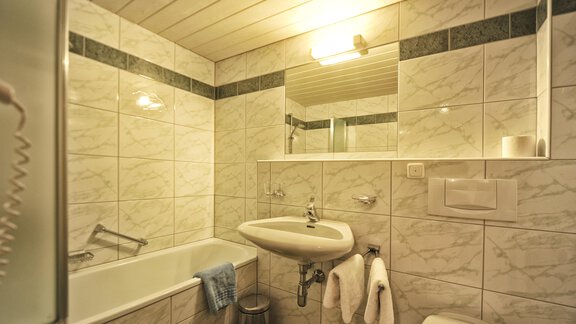 A modern bathroom with a bathtub and a sink. White tiles and soft lighting create a bright, inviting atmosphere. - Haus Timmelseck - Obergurgl 