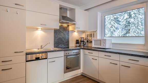 A modern designed kitchen with white cabinets and a window that looks out on snow-covered trees. The equipment includes a stove, a sink, and modern kitchen appliances. - Haus Timmelseck - Obergurgl 