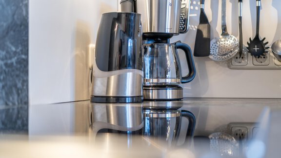 A modern kettle and a coffee machine are on a shiny kitchen countertop. In the background, various cooking utensils and an electrical outlet are visible. - Haus Timmelseck - Obergurgl 
