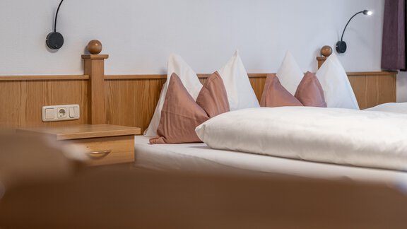 A cozy bedroom with a double bed, white bedspreads, and decorative pillows. Two reading lamps are mounted on the wall, and a nightstand stands next to it. - Haus Timmelseck - Obergurgl 