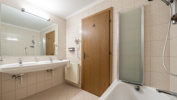 A modern bathroom with white tiles and a bathtub. There is a large mirror and a wooden interior door. - Haus Timmelseck - Obergurgl 