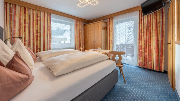 A cozy bedroom with a large bed and modern furniture. The windows are decorated with colorful curtains and there is a small table with chairs. - Haus Timmelseck - Obergurgl 