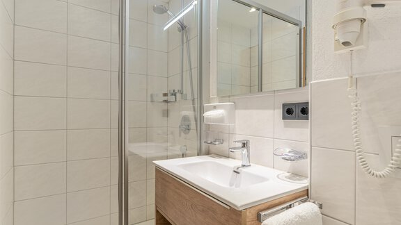 A modern bathroom with a shower and a wooden sink. The walls are tiled in light colors and there is a large mirror. - Haus Timmelseck - Obergurgl 